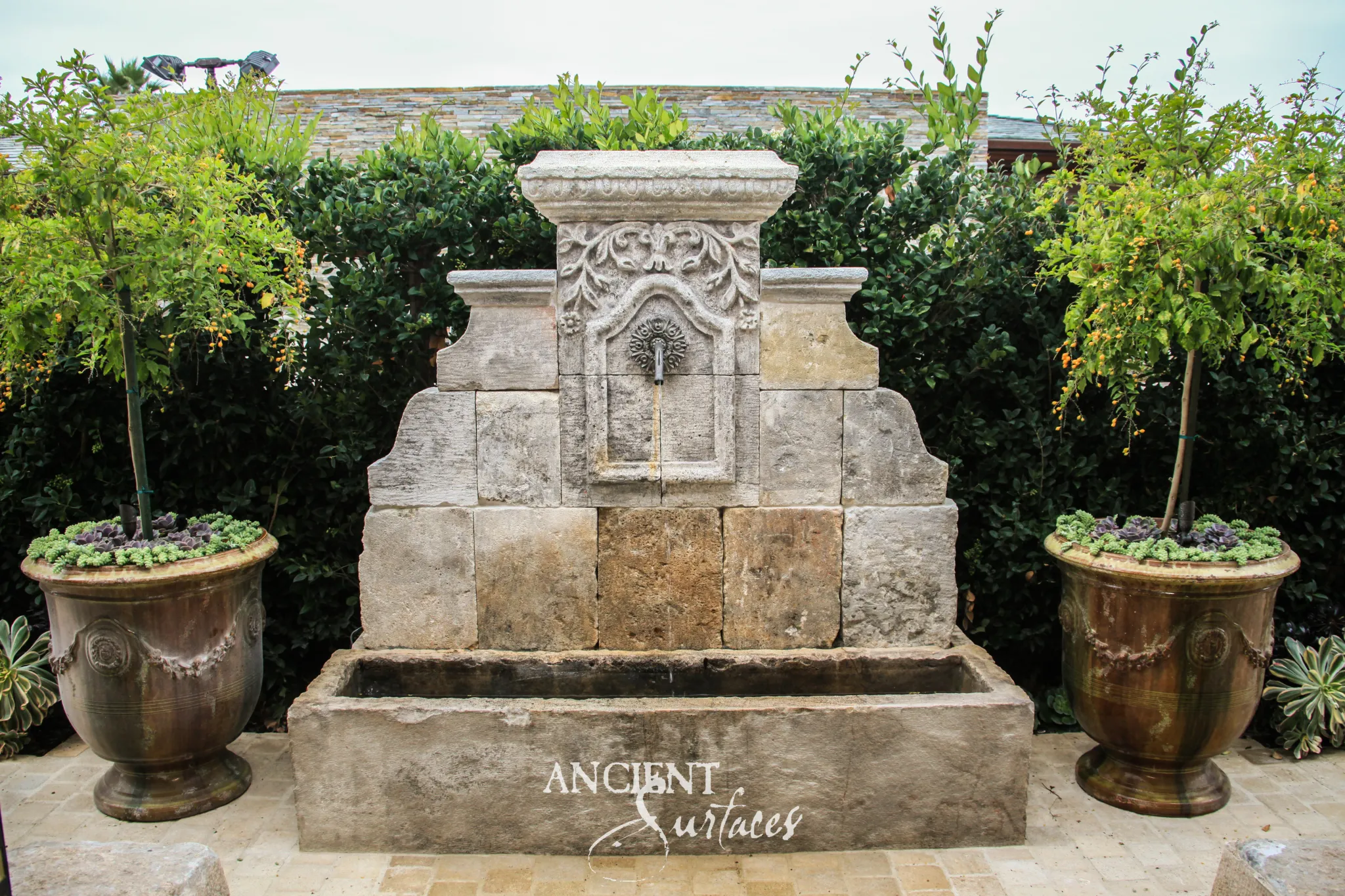Antique reclaimed limestone wall fountain flanked by glazed urns against a clipped hedge